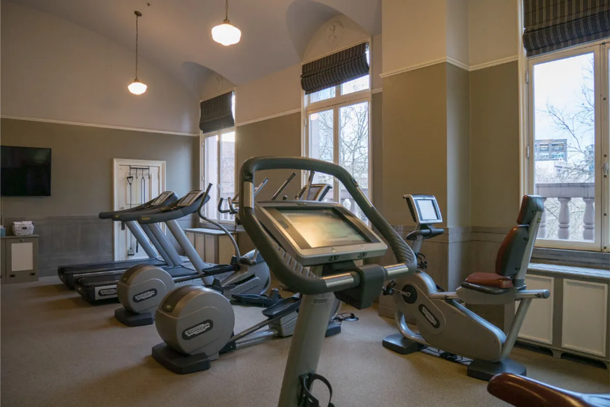 A modern fitness center featuring elliptical machines and treadmills positioned by large windows, allowing natural light to fill the room. Video screens add a contemporary touch, and the setup encourages both solo and group workouts.