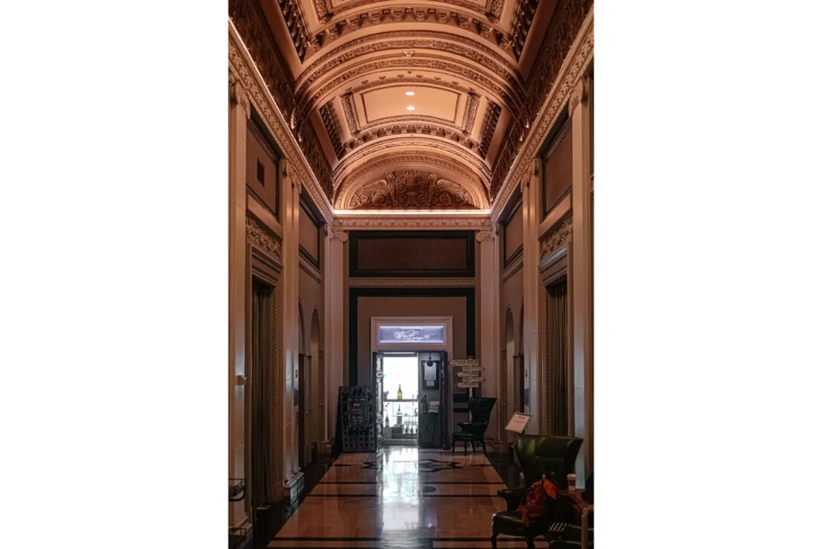 A view of the hotel's elegant double doors leading into a sophisticated, historic lobby space with ornate ceiling details, intricate moldings, and a polished floor. The openness and entrance-focused composition hint at seamless access to the outside world and the city's offerings.