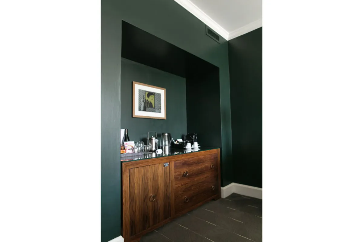A warm, intimate corner featuring a wooden cabinet, a well-equipped coffee station, and framed artwork. The atmosphere is cozy and sophisticated, ideal for enjoying a morning coffee while appreciating local art.
