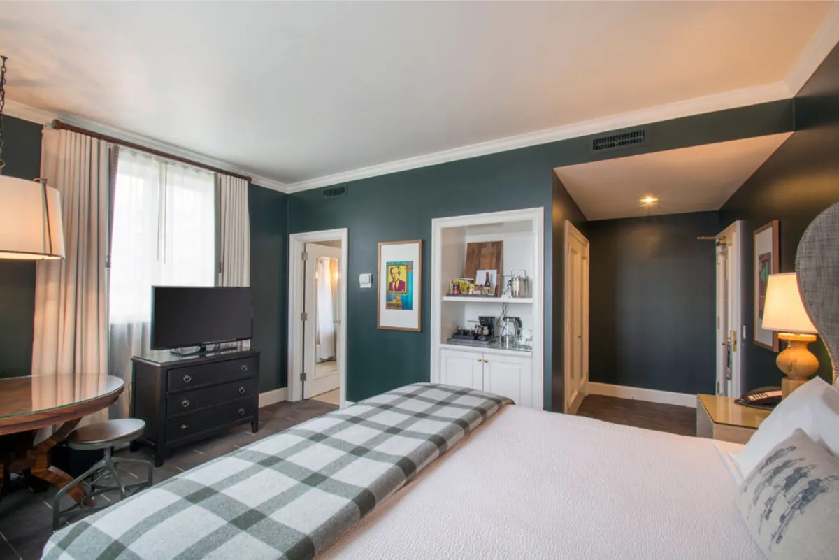 A cozy bedroom scene featuring a king-sized bed with a plaid blanket, a wooden desk and chair positioned near the window, a television mounted on the wall, and thoughtfully placed artwork. The space is bathed in soft, inviting light, evoking a sense of relaxation and comfort.