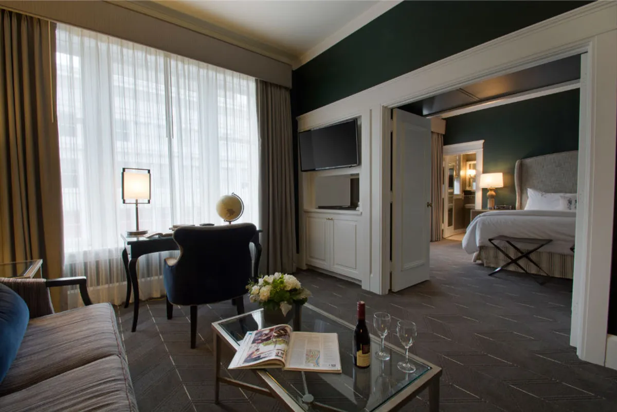 The image showcases a sophisticated hotel suite in Portland's West End, featuring a sunlit sitting area with a plush armchair beside a coffee table set with wine and a magazine. Sheer curtains filter natural light through a large window, and an inviting bedroom is visible through an open door, hinting at comfort and elegance.
