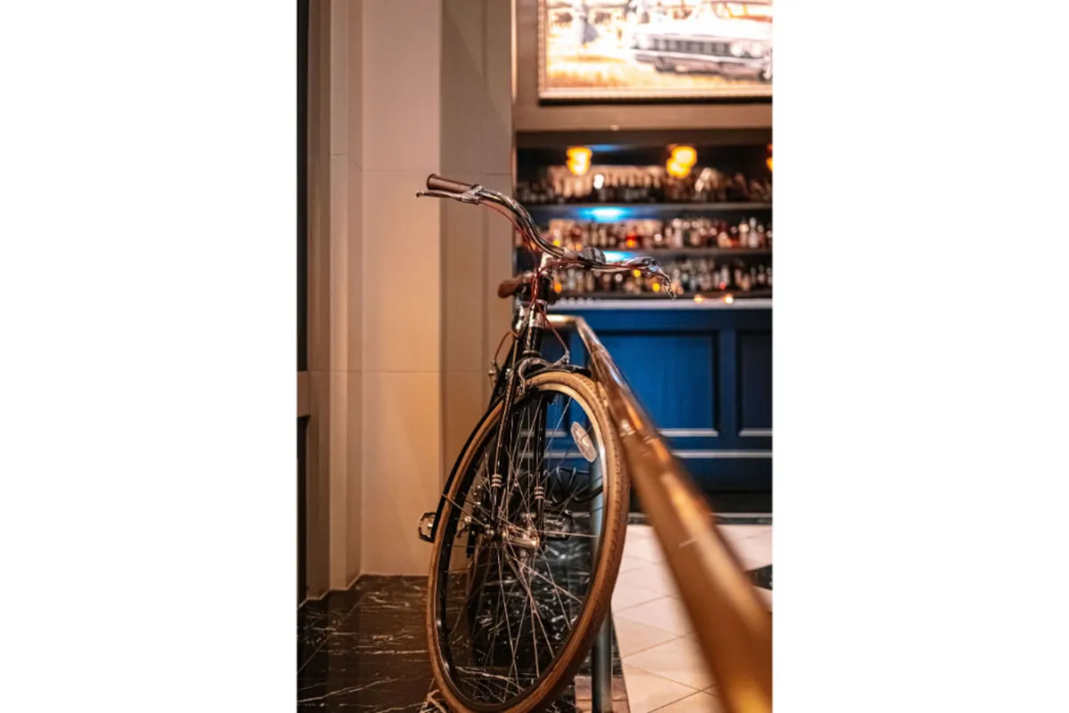 A welcoming hotel bar area with a prominent bicycle displayed near the counter, surrounded by patrons enjoying drinks and conversation under nostalgic lighting and artistic wall décor. The scene suggests both a social hub and the hotel's commitment to helping guests explore the city by bike.