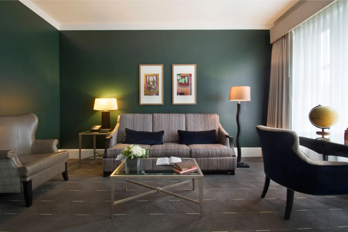 A cozy living space featuring a comfortable sofa against a green accent wall, complemented by a glass coffee table and curated artwork on the wall. The scene is awash with natural light, creating an inviting atmosphere for relaxation or socializing.